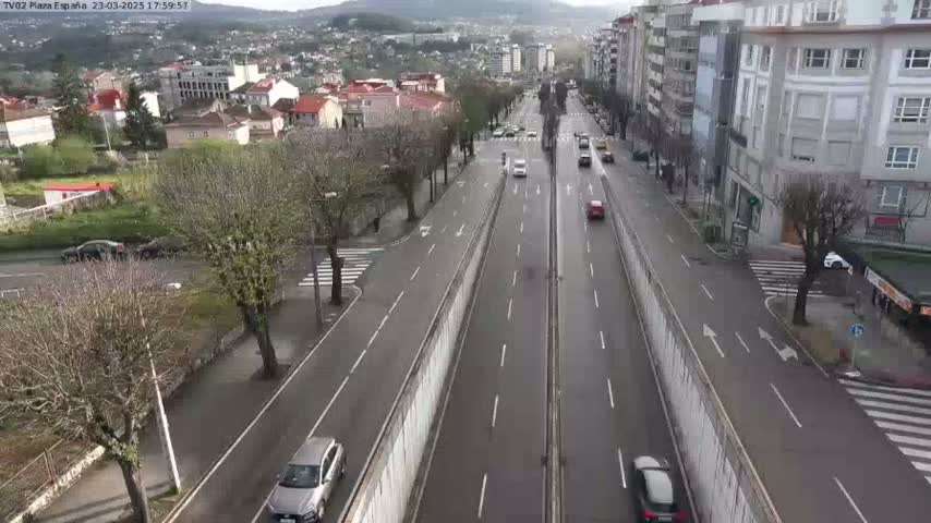 A high-angle view shows a multi-lane road with sparse traffic, flanked by buildings and trees under an overcast sky.