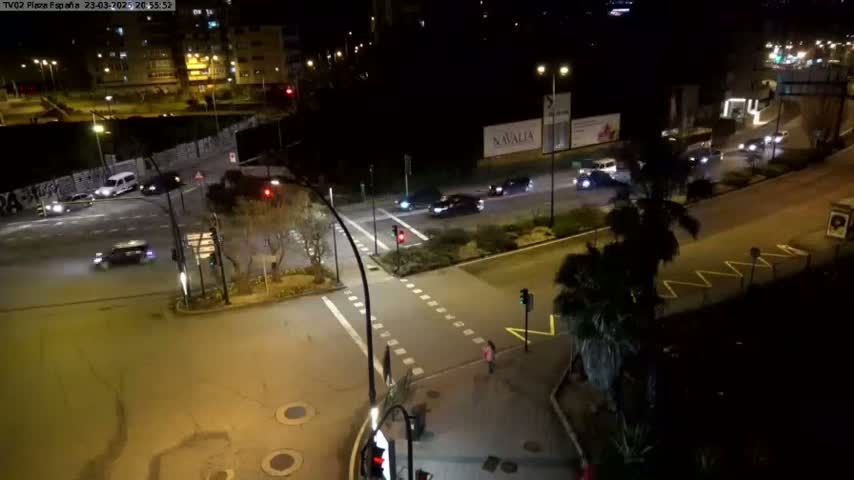 A nighttime view of a street intersection with several cars and pedestrians, illuminated by streetlights.