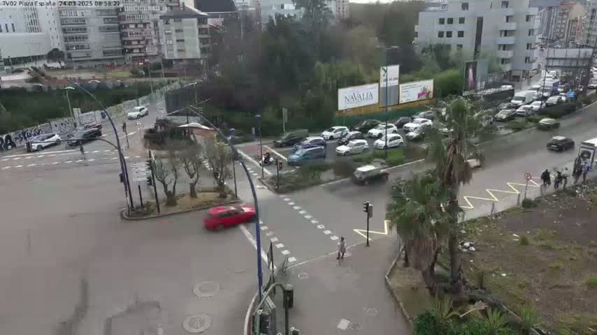 A wet city intersection shows several cars and pedestrians, with buildings and greenery in the background.