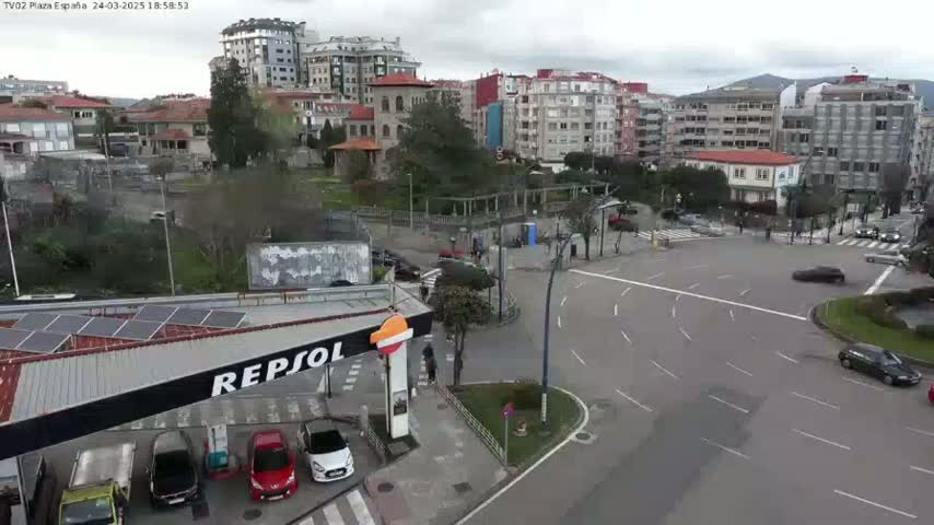 An aerial view of a city intersection on an overcast day, showing several cars, a gas station, and buildings surrounding a park-like area.
