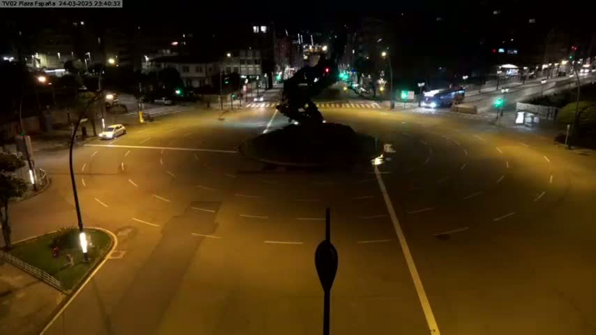 A nighttime aerial view of a mostly empty roundabout with a few vehicles and buildings in the background under well-lit conditions.
