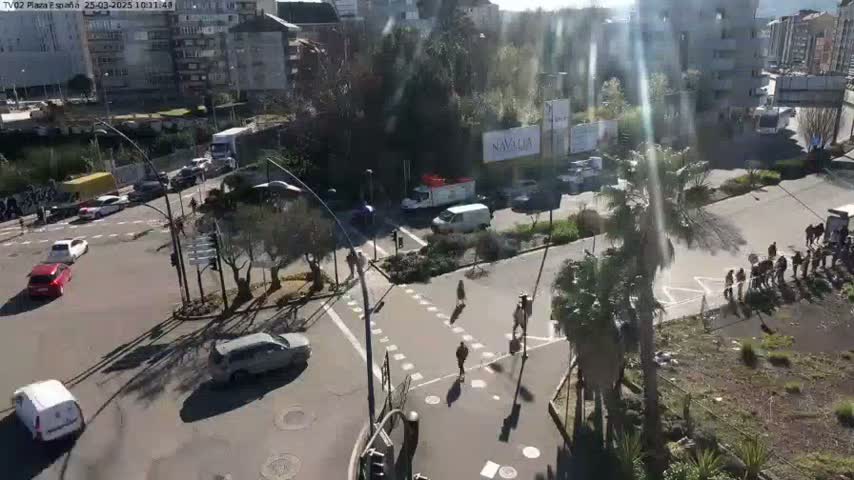 A sunny day overlooking a city intersection with several pedestrians and cars, some palm trees, and buildings in the background.