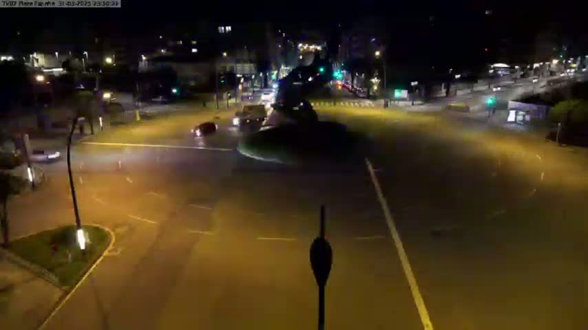 A nighttime aerial view shows a wet, dimly lit intersection with several vehicles moving through it.