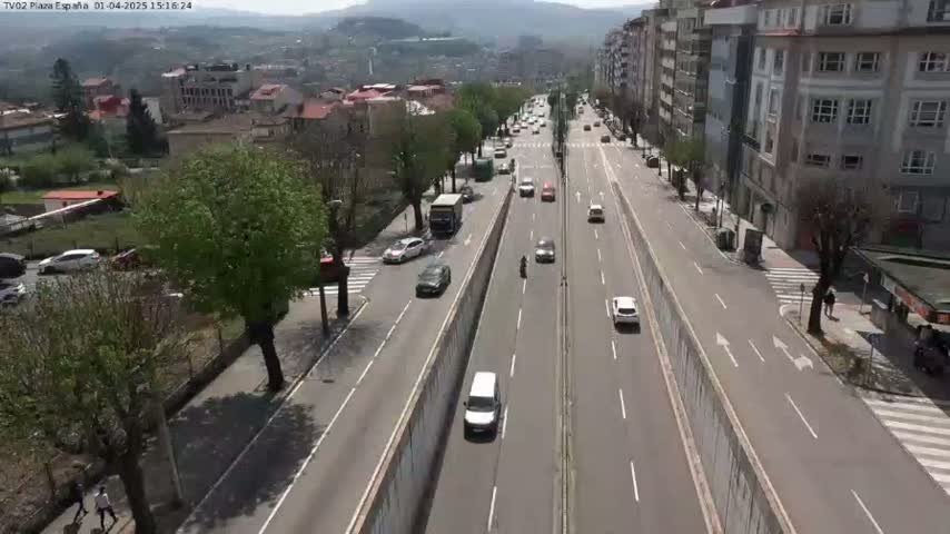 A sunny day reveals a multi-lane highway cutting through a town, with several cars and a bus traveling in both directions, surrounded by buildings and lush green trees.