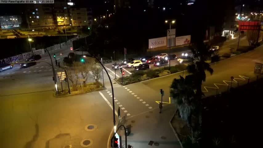 A nighttime aerial view of a city intersection with several cars stopped at traffic signals, surrounded by buildings and palm trees.