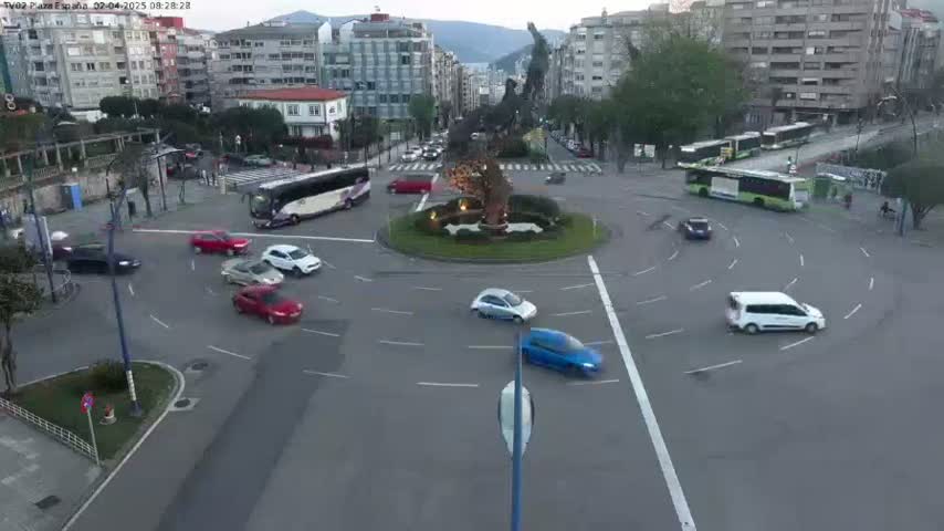 Several cars navigate a roundabout in a city setting under overcast skies.