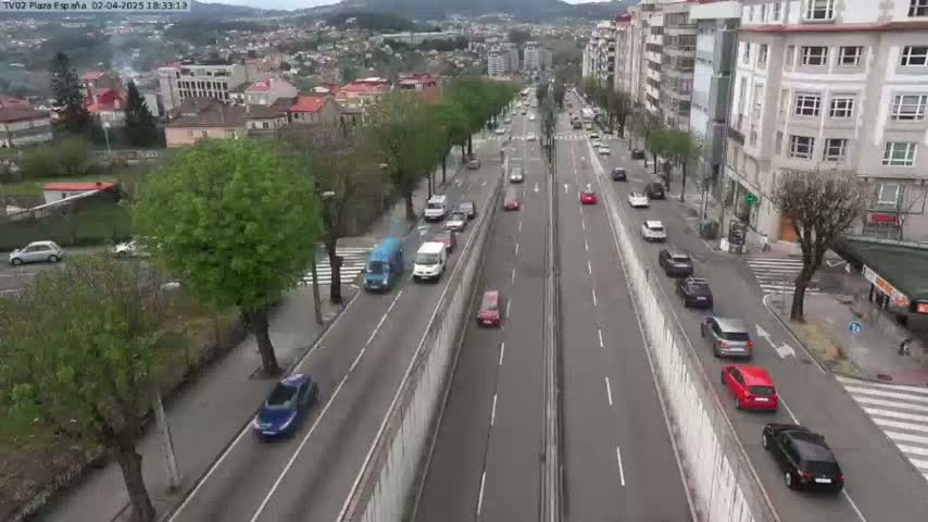 A high-angle view of a multi-lane road with moderate traffic, surrounded by buildings and lush greenery under a partly cloudy sky.