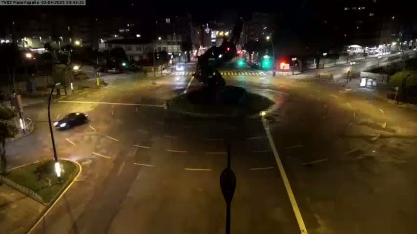 A wet, nighttime roundabout with a dark sculpture in the center, illuminated by streetlights, with a few cars sparsely visible.