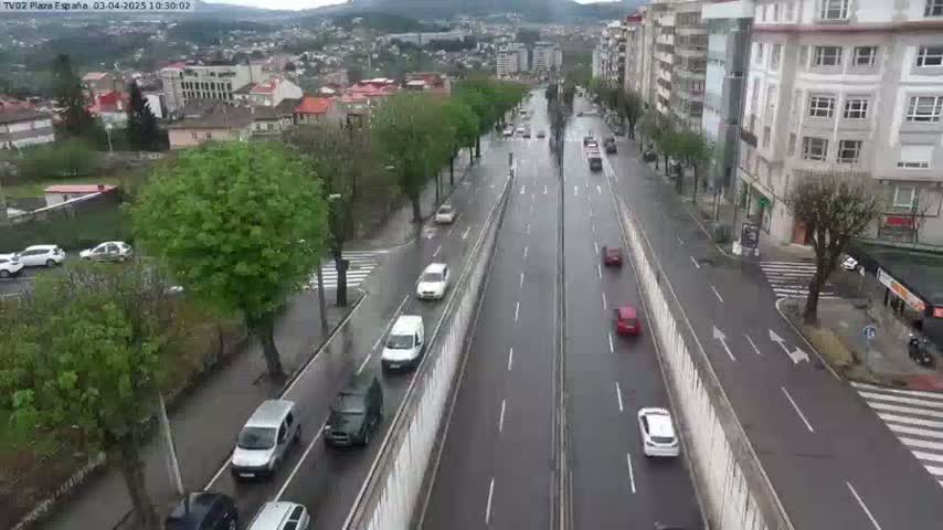A rain-slicked multi-lane road with several cars driving on it, surrounded by buildings and lush green trees.