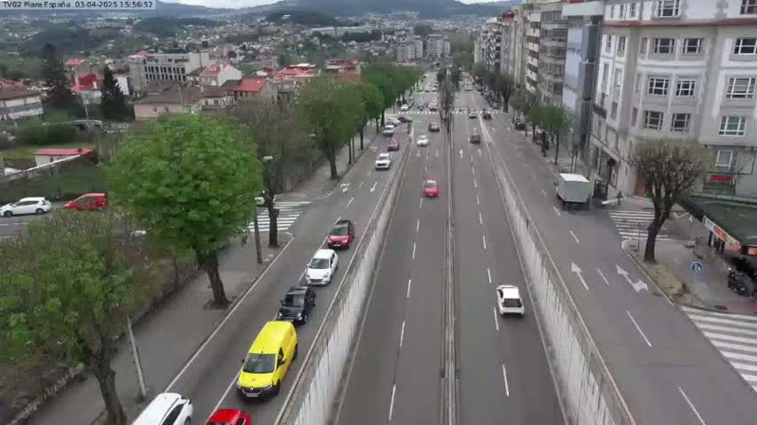 A city street scene shows several cars driving on a divided roadway, lined with buildings and trees under a mostly cloudy sky.