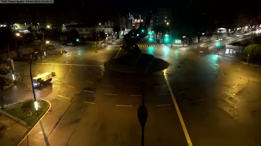 A wet, empty roundabout at night, illuminated by streetlights, with a few vehicles visible in the distance.