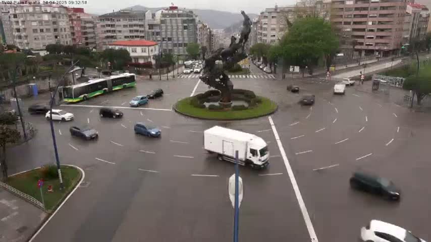 A wet roundabout with several cars and a bus, centered around a large sculpture, is surrounded by buildings under an overcast sky.