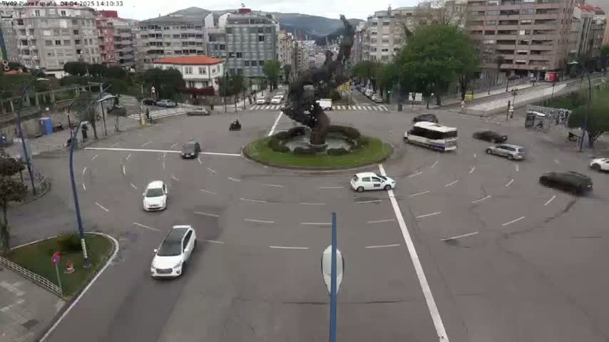 A roundabout with several cars and a bus in a city, under an overcast sky.