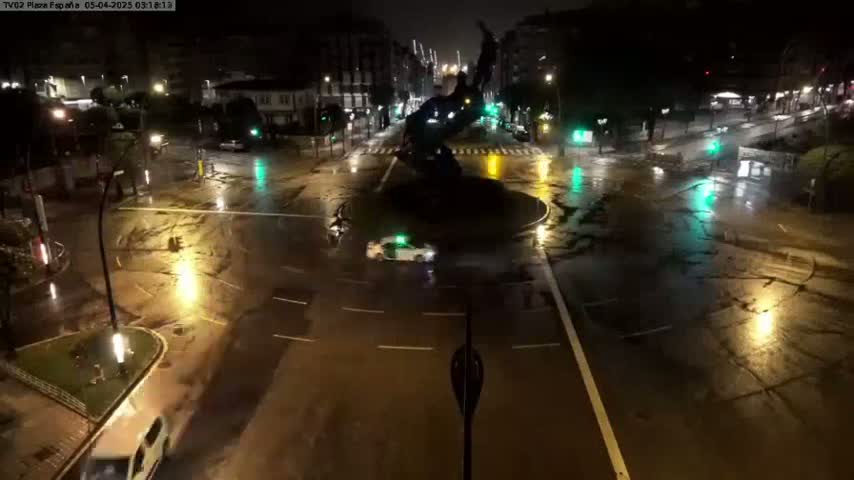 A wet, nighttime street scene shows a roundabout with a dark statue in the center, several cars, and streetlights reflecting on the puddles.