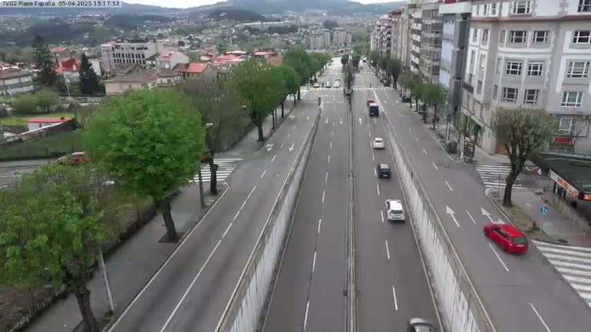 A mostly empty multi-lane road cuts through a city, lined with buildings and trees under a clear sky.
