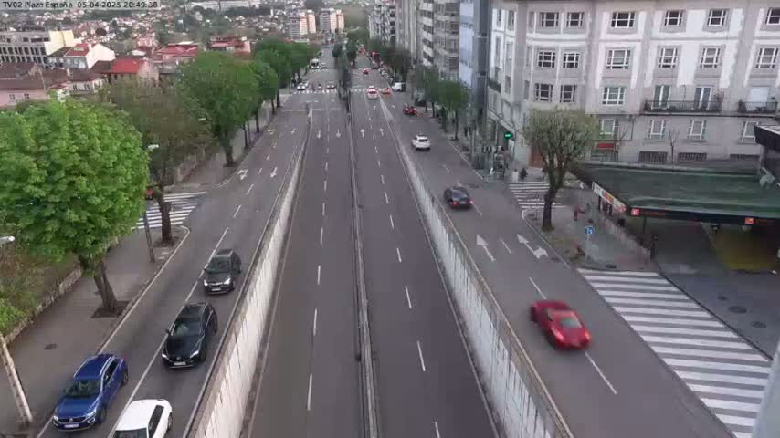 A multi-lane road with several cars driving on it is flanked by buildings and trees under a daytime sky.
