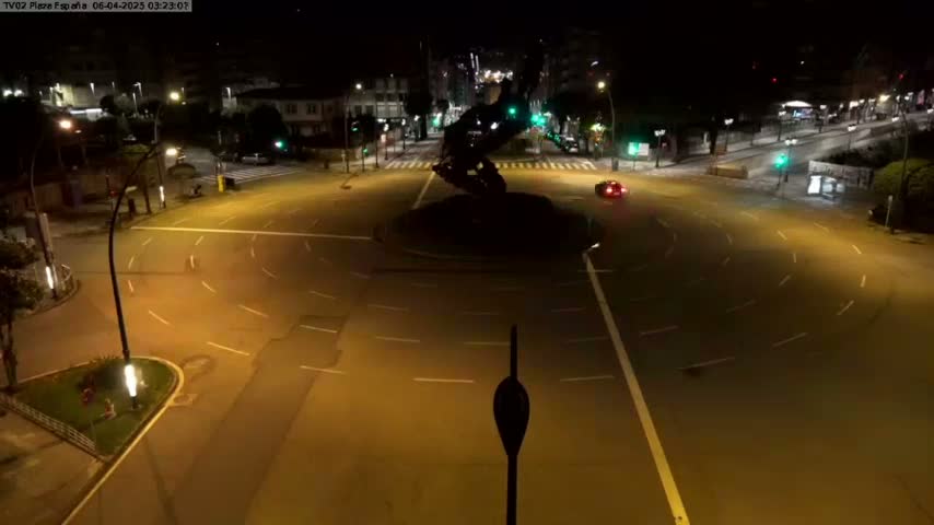 A dark, nighttime aerial view of a roundabout with a large, dark sculpture at its center, illuminated by streetlights, with one car visible.