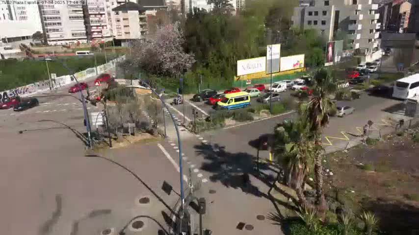 A sunny day shows a street intersection with several cars and a bus, surrounded by buildings, trees, and palm trees.