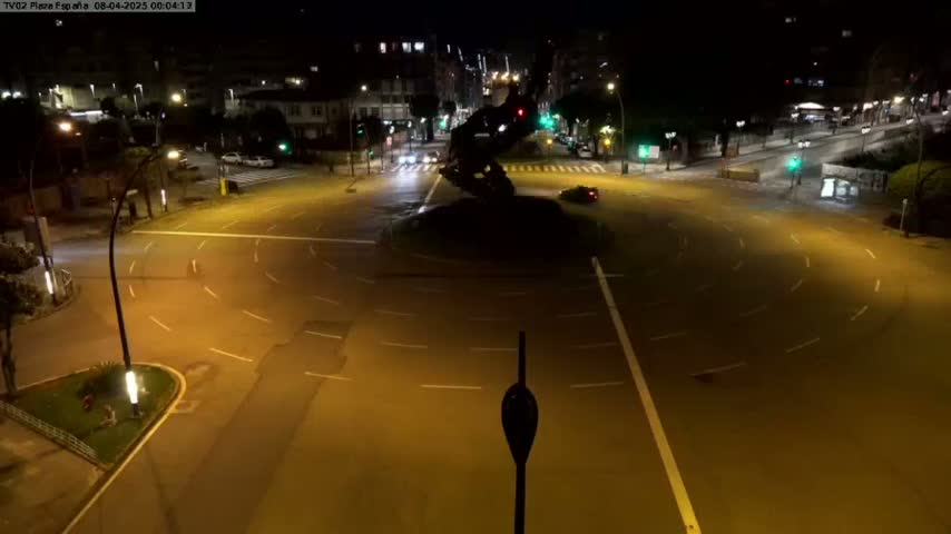 A nighttime aerial view shows a mostly empty roundabout with a dark statue or sculpture in the center, illuminated by streetlights.