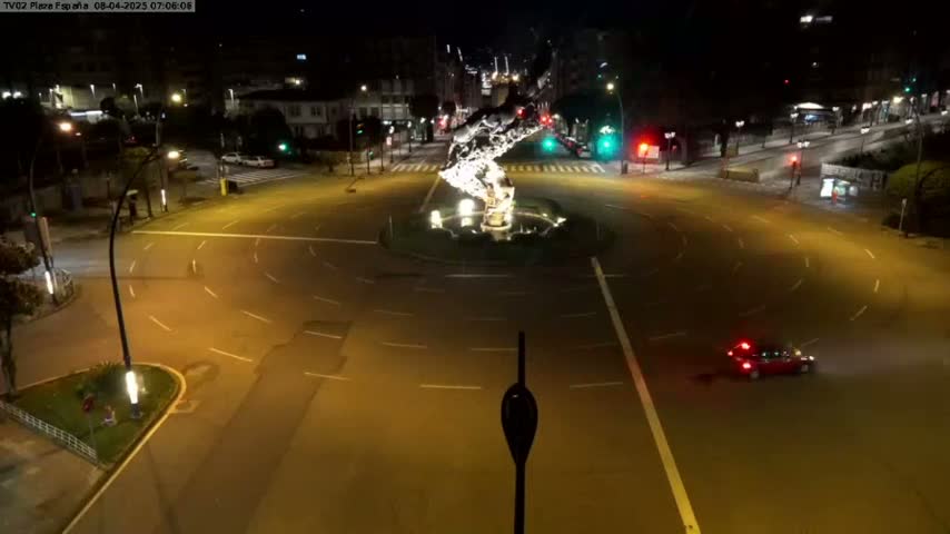 A nighttime aerial view of a roundabout with a central illuminated sculpture, with a single car visible on the road.