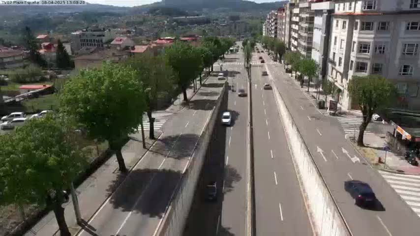 A sunny day view of a multi-lane road bordered by buildings and trees, with several cars driving in both directions.