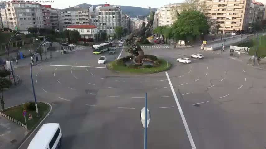 A roundabout with several cars and a large, abstract sculpture in the center, under a mostly clear sky.