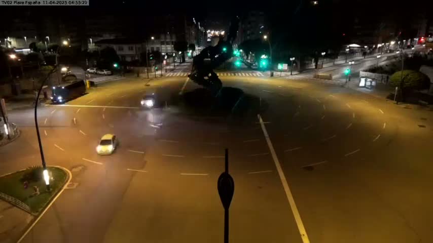 A nighttime, overhead view of a roundabout with a few cars and a large dark sculpture in the center.