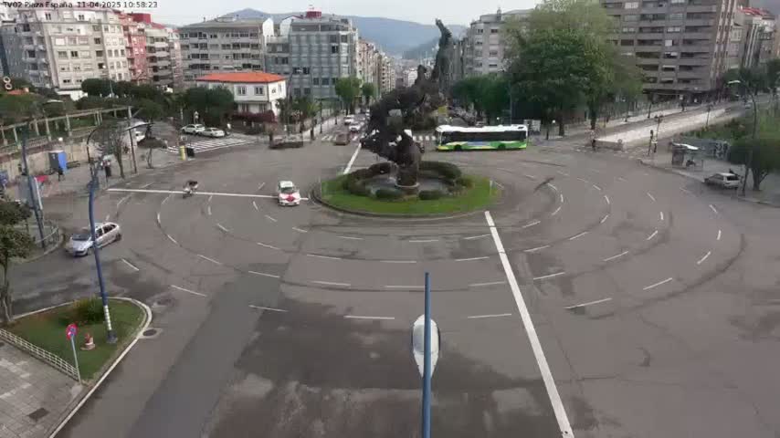 A roundabout with a large dark statue at its center is surrounded by buildings and a few cars and a bus on an overcast day.
