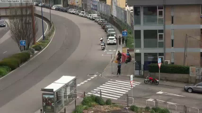 A curved road with parked cars and several people standing near motorcycles and a red scooter, next to a multi-story building under a bright, sunny sky.