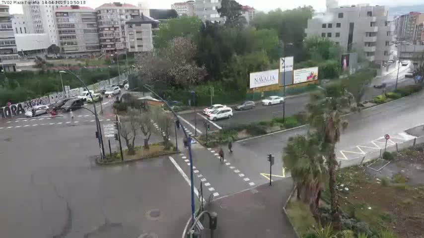 A wet, city intersection with several cars and pedestrians, surrounded by buildings and trees in rainy weather.