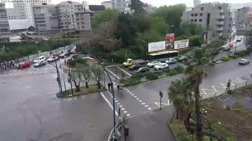A rainy city intersection shows several cars stopped at a crosswalk where pedestrians are crossing, with buildings and trees visible in the background.