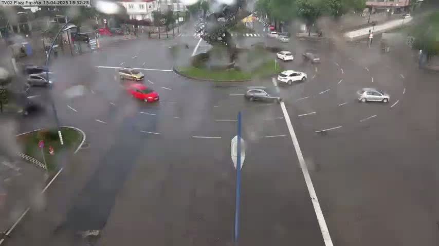 Several cars navigate a rain-slick roundabout in a city setting.