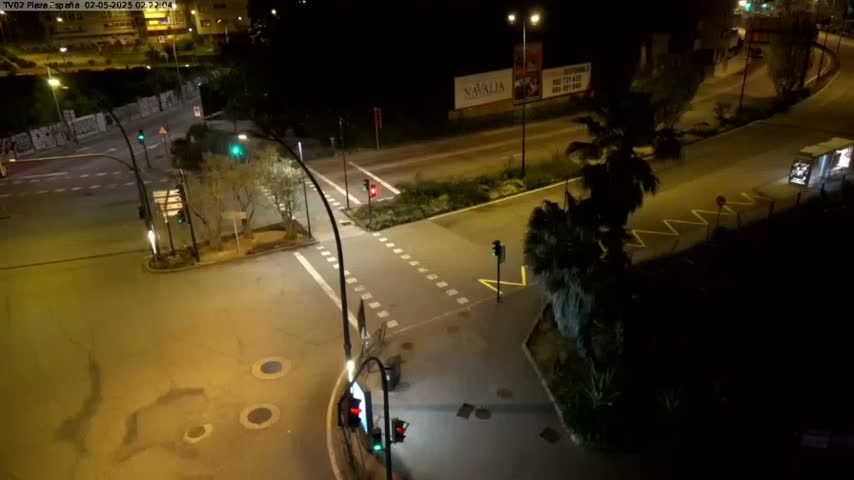 A nighttime, high-angle view of a street intersection with traffic signals, sparse vehicles, and surrounding vegetation.