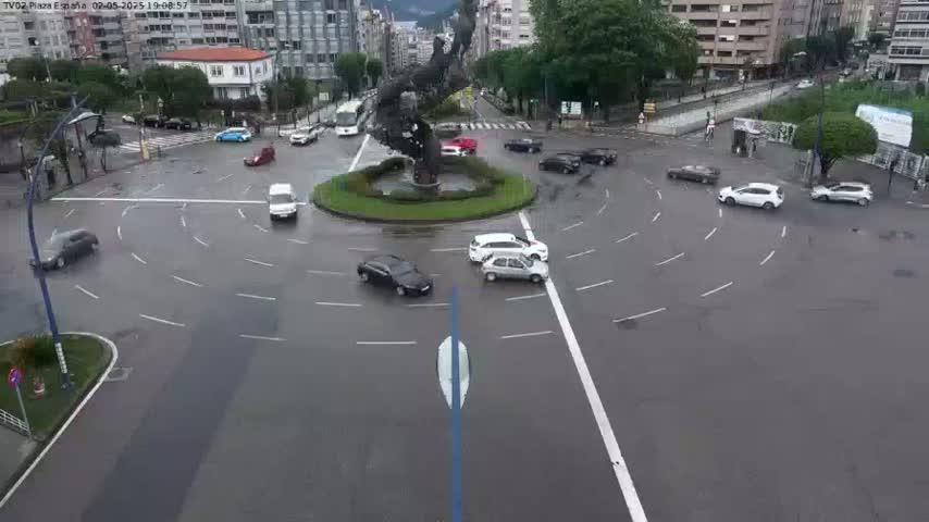 Several cars navigate a wet roundabout centered around a statue, with surrounding buildings visible in the rainy weather.