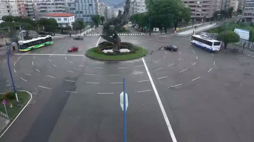 A wet roundabout with several cars and buses is centered around a sculpture.