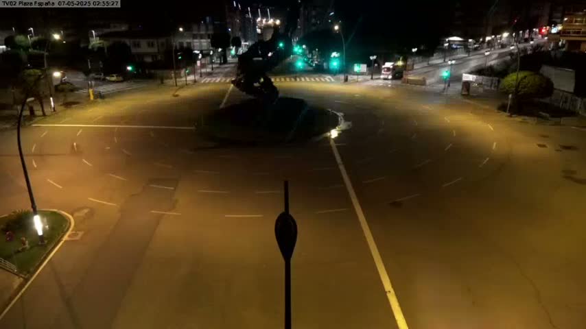 A dark, empty roundabout at night, illuminated by streetlights, with a large sculpture in its center.