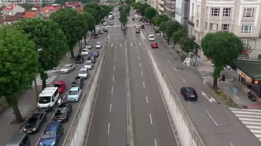 A high-angle view shows a multi-lane road with cars driving in both directions, bordered by green trees and buildings, under a seemingly clear sky.