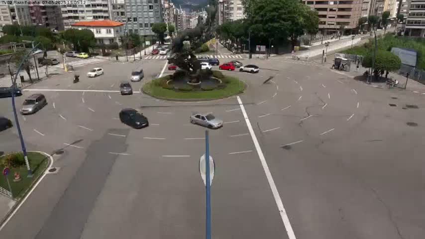 A sunny day shows several cars navigating a roundabout centered around a large sculpture, with buildings and trees visible in the background.