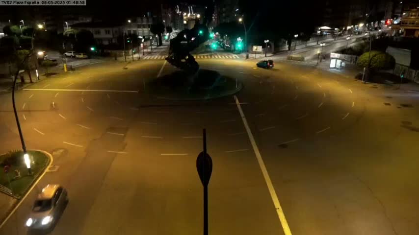 A nighttime, high-angle view shows a mostly empty roundabout with a dark statue at its center, illuminated by streetlights.