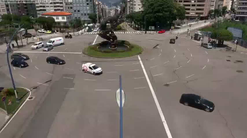 A high-angle view of a roundabout with several cars, a large sculpture in the center, and buildings visible in the background on a sunny day.