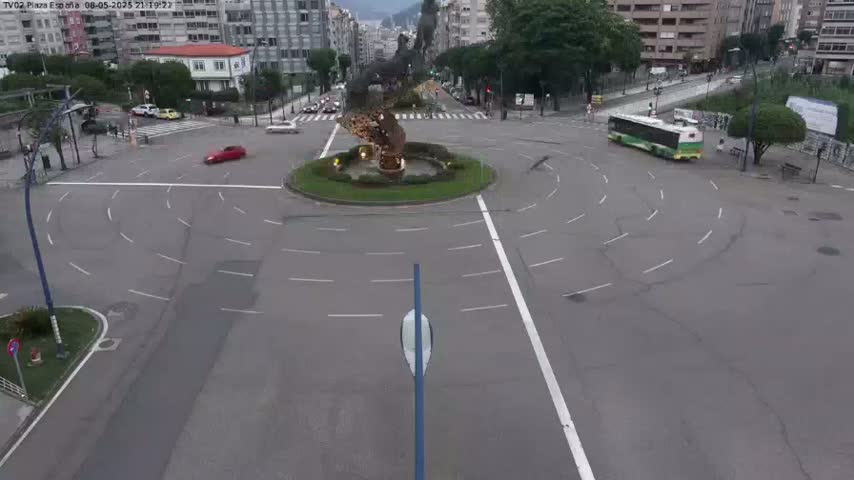 A mostly empty roundabout with a central fountain and a few cars and a bus is visible in an overcast daytime scene.