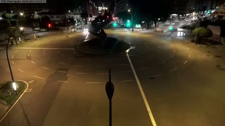 A nighttime, high-angle view of a mostly empty roundabout with a dark sculpture in the center, surrounded by streets with sparsely driving cars and illuminated by streetlights.