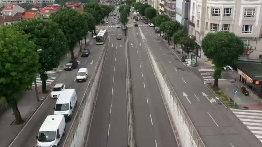 A multi-lane roadway with light traffic, flanked by buildings and trees under a partly cloudy sky.