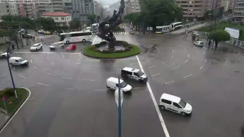 Several cars navigate a rain-slick roundabout centered on a large, dark statue, with buses and other vehicles visible in the background.