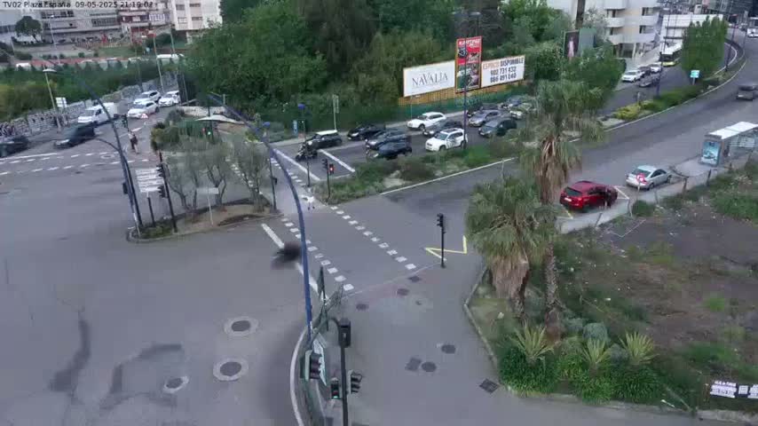 A high-angle, daytime view shows a multi-lane intersection with several cars, pedestrians, and palm trees under a partly cloudy sky.