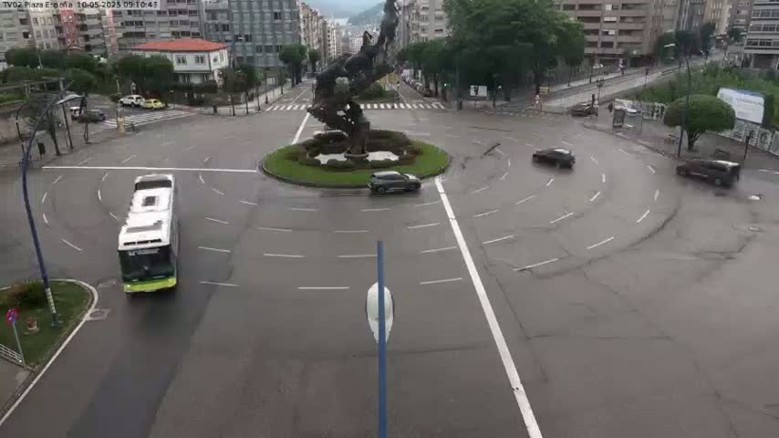 A wet roundabout with several cars and a bus, centered around a large statue.