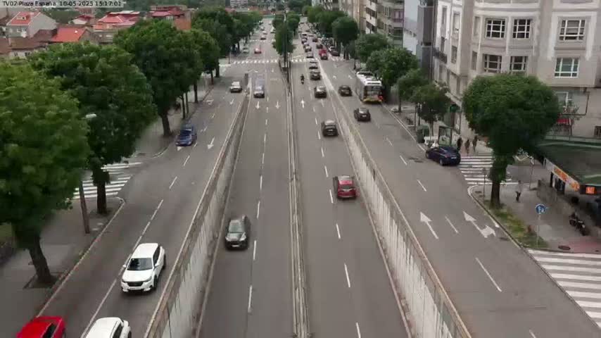 A multi-lane road with moderate traffic flows through a city with buildings lining the sides under an overcast sky.