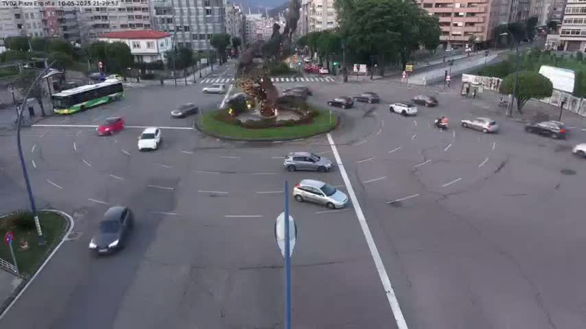 A roundabout with several cars and a bus in a city setting under twilight conditions.