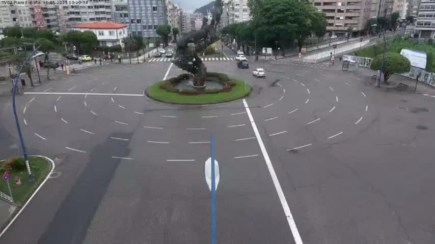 A roundabout with a large sculpture in the center, surrounded by buildings and a few cars, on a wet, overcast day.