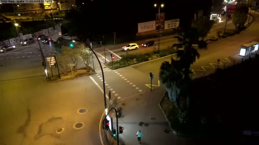 At night, a few cars are driving through a well-lit intersection surrounded by trees and buildings.